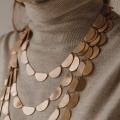 Dekolt kobiety ubranej w beżowy golf i 4 złocone naszyjniki projektu Marty Szafraniec, różnej długości. Jeden, najkrótszy, rozmiaru XS, zawieszony tuż przy szyi. Każdy następny dłuższy o kilka elementów czyli połówek koła, z których jest wykonany.
