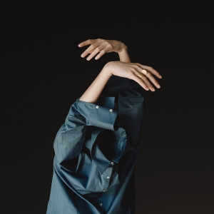 Wystające ręce z koszuli niebieskiej,  uniesione w górę. Dłonie, skierowane na siebie, jak wiszące liście monstery. Na palcu duży, otwarty, złocony pierścionek Halfring, marki Saffa z Katowic.