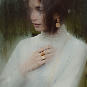 Piękna, zamyślona kobieta za zaparowaną szybą, w białym golfie, w złoconych kolczykach z koła i połówki. Z dłonią na serca. Na palcach dwa otwarte, złocone pierścionki, marki Saffa, z nowej kolekcji Halfring.