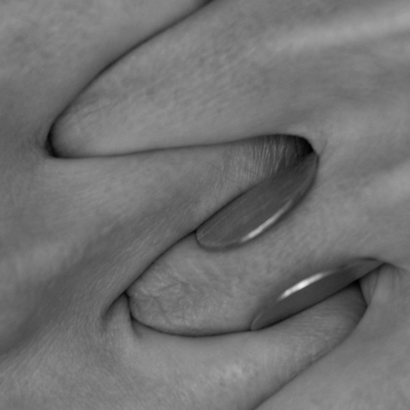 Detal. Wystające na sztorc, z nad palców splecionych ciasno razem, krawędzie srebrnego, otwartego pierścionka Halfring, pomysłu Marty Szafraniec. Duży, symetryczny, wyszczotkowany na powierzchni i wybłyszczony na krawędziach. Piękny.