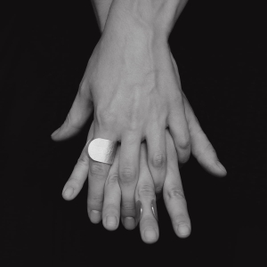 Przykryte sobą dłonie kobiece, jedna na drugą, w geście jedności i bliskości, na jednym palcu srebrny pierścionek z szeroką, otwartą obrączką, wyszczotkowany na powierzchni i wybłyszczony na krawędziach. Zadbany detal w stylu marki Saffa z Katowic.