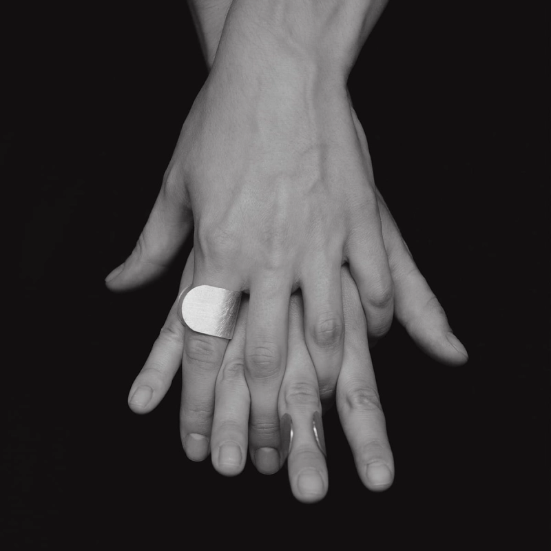 Przykryte sobą dłonie kobiece, jedna na drugą, w geście jedności i bliskości, na jednym palcu srebrny pierścionek z szeroką, otwartą obrączką, wyszczotkowany na powierzchni i wybłyszczony na krawędziach. Zadbany detal w stylu marki Saffa z Katowic.