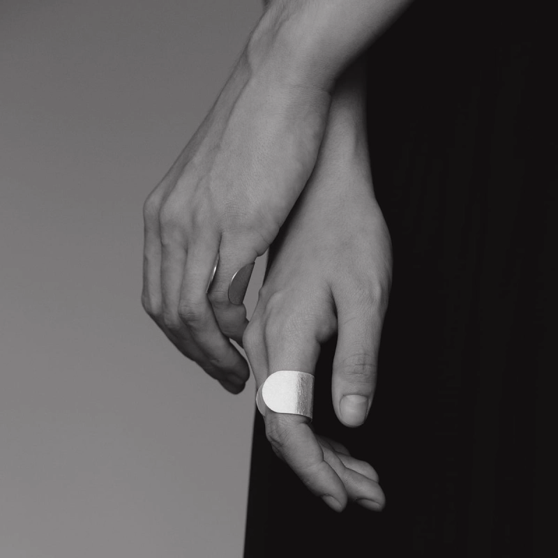 Luźno puszczone dłonie,na czarnym tle, na 2 palcach srebrne pierścionki z najnowszej kolekcji Halfring, wykonane ręcznie w trudnej do znalezienia pracowni projektowania biżuterii Saffa w Katowicach. Lśniąca, duża, otwarta, unikatowa biżuteria.