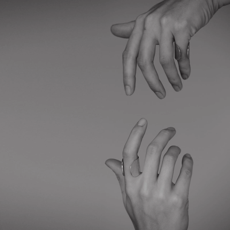 Dłonie spoglądające na siebie w pionie. Na jednym palcu srebrny pierścionek z najnowszej kolekcji Halfring, duży, otwarty, z szeroką obrączką, wyszczotkowany na powierzchni i wypolerowany na krawędziach. Minimalistyczny w stylu marki Saffa.