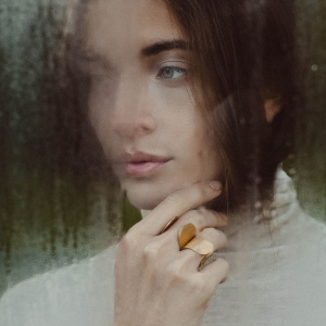 Dwa złocone, duże, otwarte pierścionki, na palcach pięknej zamyślonej kobiety, z dłonią przy krawędzi twarzy, wybłyszczone na rantach, lśniące, projektu Marty Szafraniec, projektantki ze Śląska, w miejscu, które warto odwiedzić.