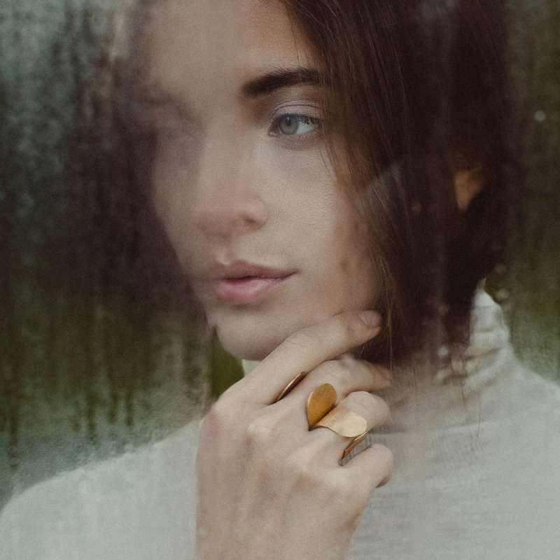 Dwa złocone, duże, otwarte pierścionki, na palcach pięknej zamyślonej kobiety, z dłonią przy krawędzi twarzy, wybłyszczone na rantach, lśniące, projektu Marty Szafraniec, projektantki ze Śląska, w miejscu, które warto odwiedzić.