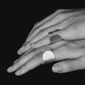 Dłonie, jedna przy drugiej, na jednym palcu każdej dłoni, srebrny pierścionek z kolekcji Halfring, duży, otwarty, minimalistyczny, w stylu marki Saffa, pracowni z Katowic z autorską biżuterią. Warte odwiedzenia miejsce na mapie Śląska.