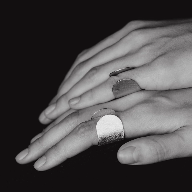 Dłonie, jedna przy drugiej, na jednym palcu każdej dłoni, srebrny pierścionek z kolekcji Halfring, duży, otwarty, minimalistyczny, w stylu marki Saffa, pracowni z Katowic z autorską biżuterią. Warte odwiedzenia miejsce na mapie Śląska.