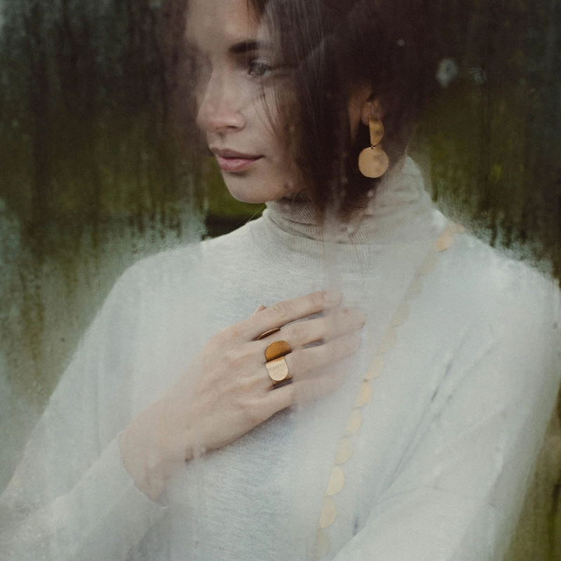 Kobieta za zaparowanym szkle, w jasnym golfie, w złoconych kolczykach i w dwóch złoconych, otwartych, minimalistycznych, unikatowych pierścionkach, biżuterii autorskiej projektu Marty Szafraniec, artystki ze Śląska, z Katowic.