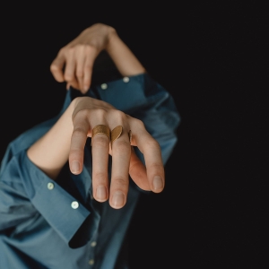 Dłonie kobiece, zabawnie wystające z nie do końca ubranej niebieskiej koszuli, na palcach 2 złocone pierścionki z kolekcji Halfring, jeden otwarty, sterczący wybłyszczonymi krawędziami w górę, drugi założony na odwrót, z szeroką obrączką.