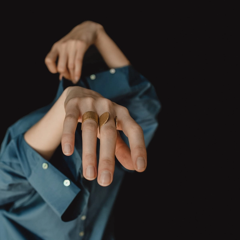 Dłonie kobiece, zabawnie wystające z nie do końca ubranej niebieskiej koszuli, na palcach 2 złocone pierścionki z kolekcji Halfring, jeden otwarty, sterczący wybłyszczonymi krawędziami w górę, drugi założony na odwrót, z szeroką obrączką.